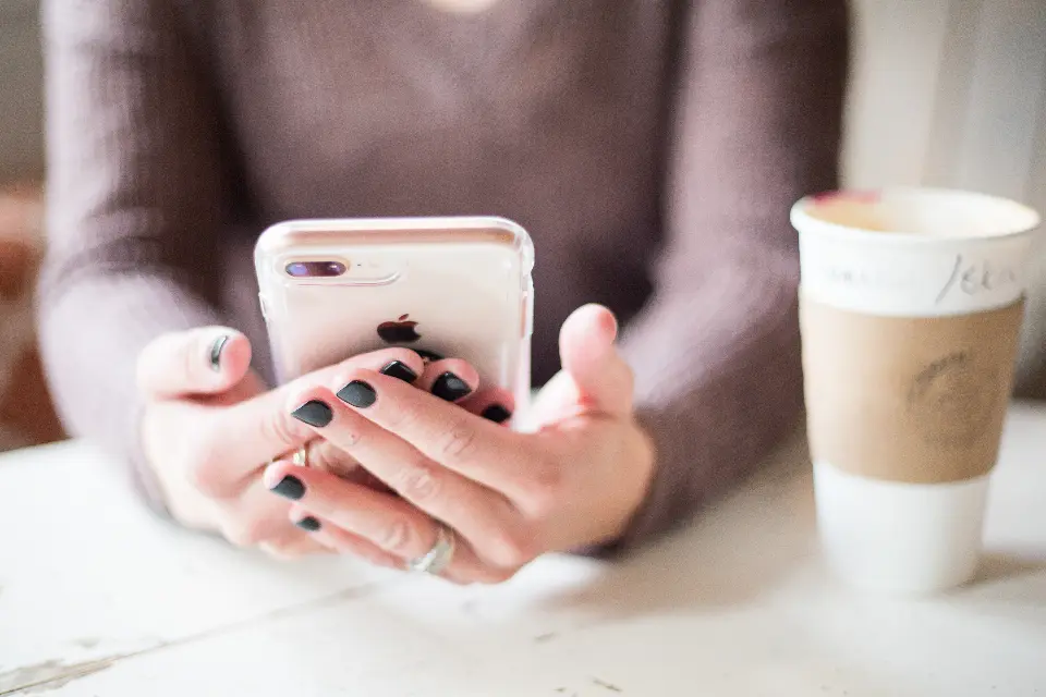 Mãos a segurar um telemóvel junto de um copo de café numa mesa clara